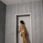 A woman stands in the doorway of a room, looking out with a neutral expression.