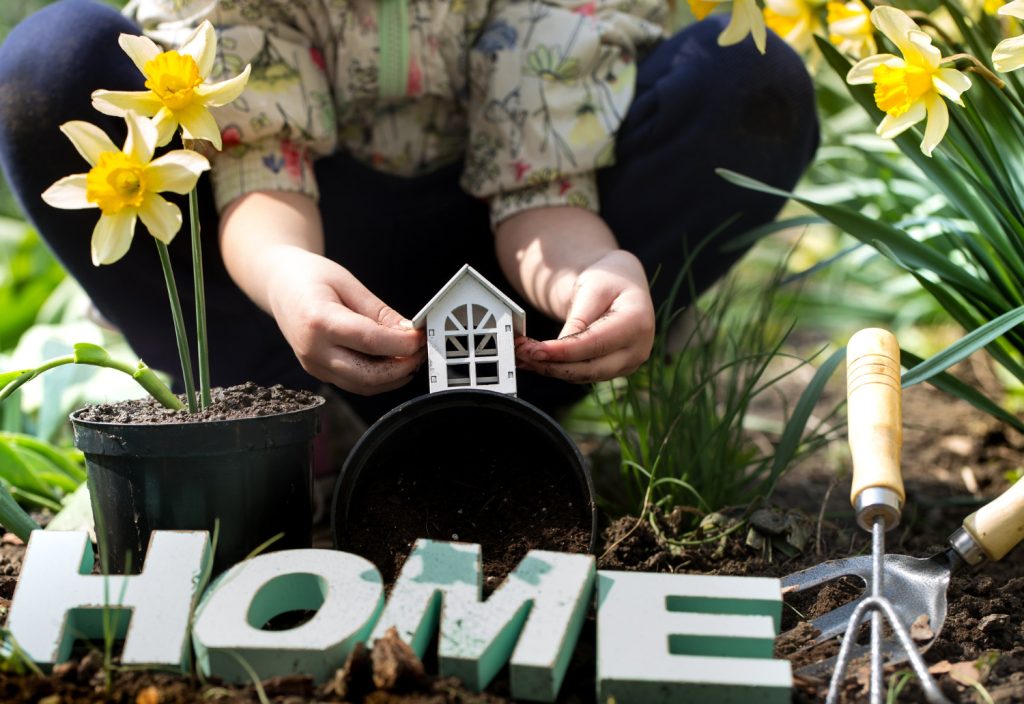A woman plants flowers in her garden, with a small house and a garden tool visible in the background