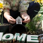 A woman plants flowers in her garden, with a small house and a garden tool visible in the background