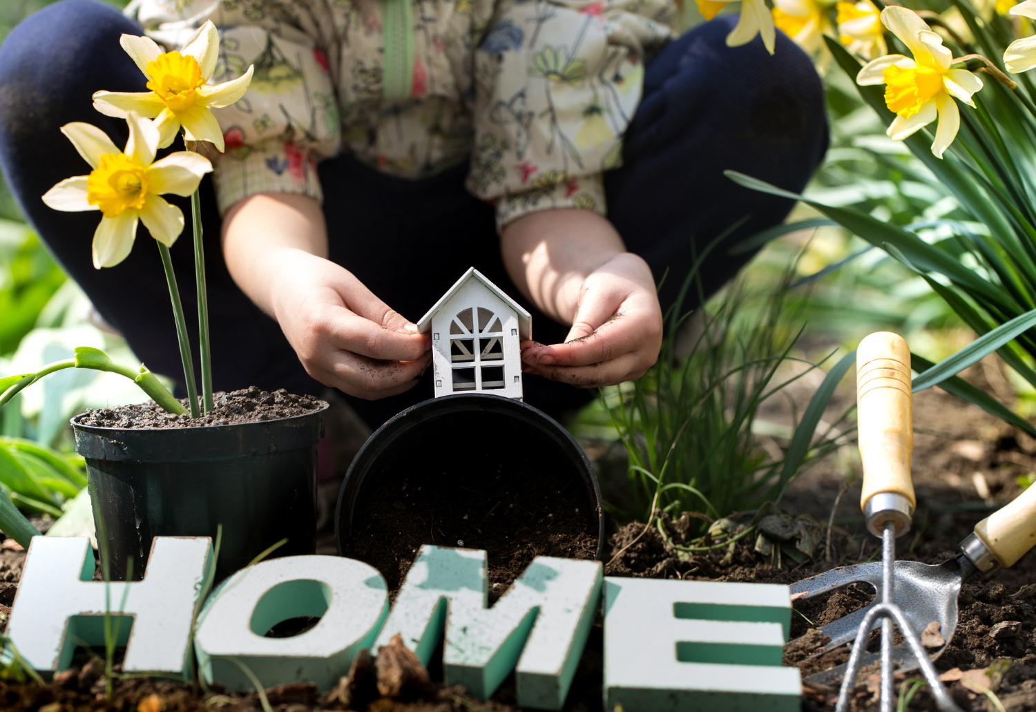 A woman plants flowers in her garden, with a small house and a garden tool visible in the background